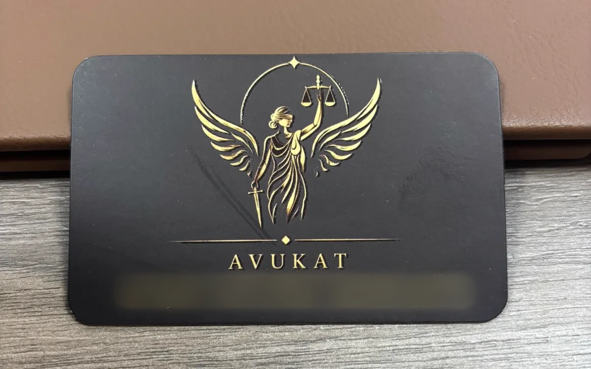 Av. A. M. A. kartvizitinin basılı hâli, siyah zemin üzerine altın kanatlı adalet figürü
